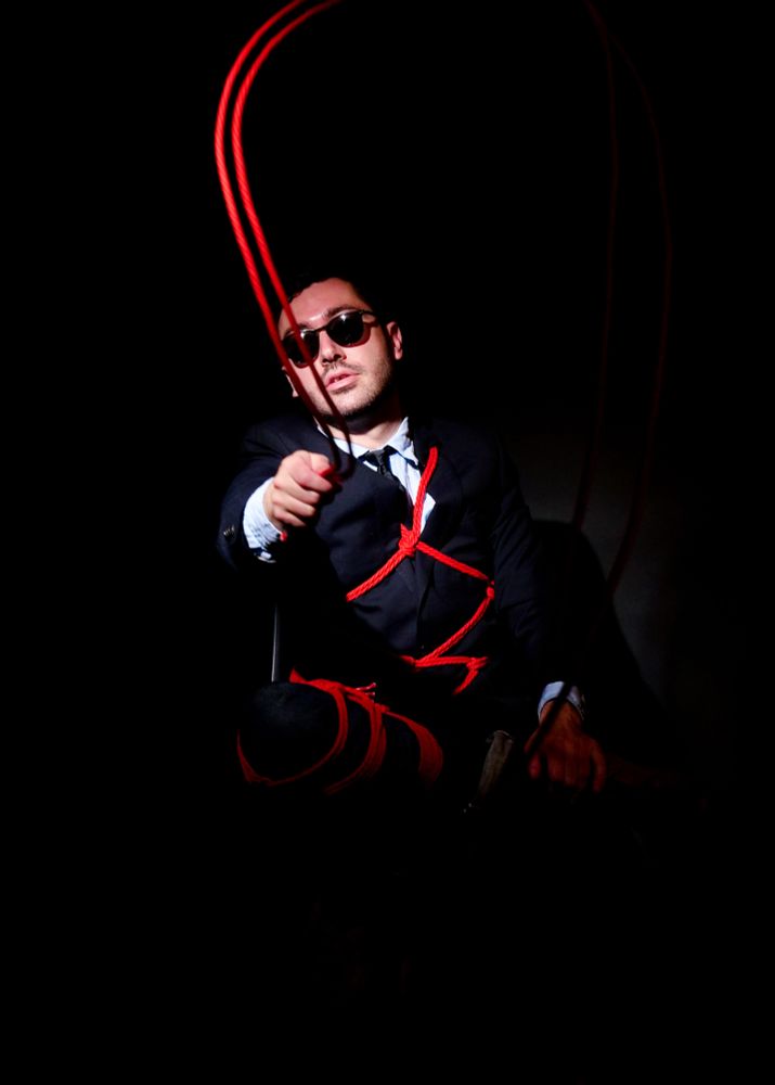 Photograph of a man wearing a black suit and tie, with red ropes, tied with Japanese rope bondage or Shibari