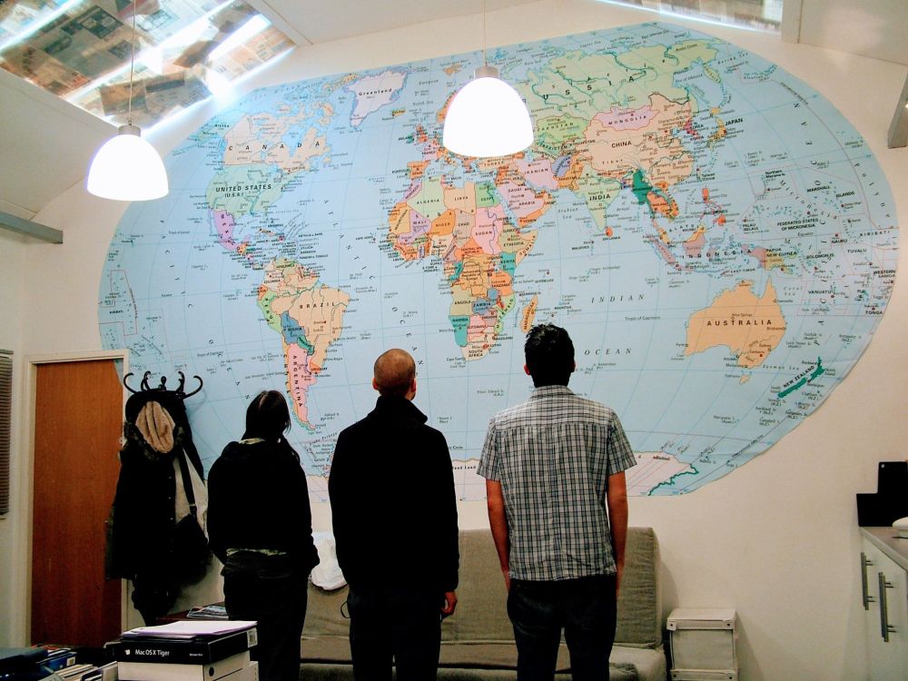 In a bright office with white walls and skylights covered in newspaper, three people in ascending height order (Katie, Kris, and James) stand with their backs to the camera. They’re looking up at a tall, wide wall. On that wall is huge map of the world.