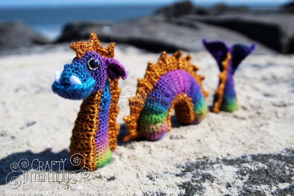 A colorful crocheted sea serpent made with rainbow and gold yarn sections is posed on a sandy beach. The serpent appears to be submerged, with its head, mid-body, and tail emerging from the sand, giving the illusion of swimming through it. Rocks and the ocean are visible in the background.