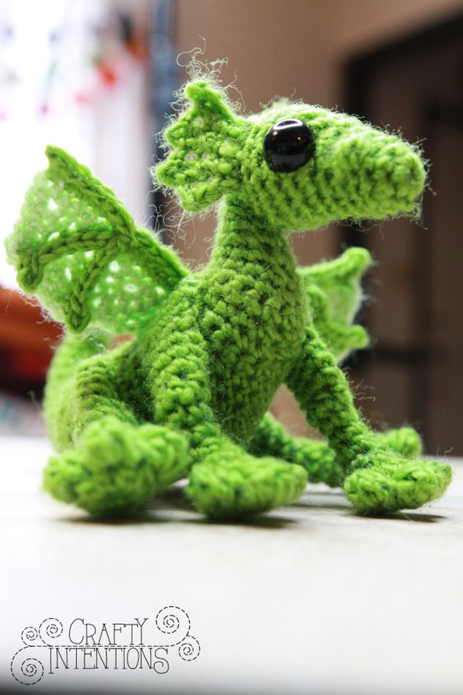 A close-up of a crocheted bright green baby dragon sitting on a light surface. The dragon has shiny black safety eyes, small wings with openwork stitching, and a sweet, slightly fuzzy texture.