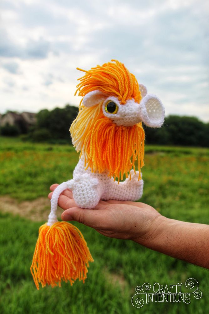 A crocheted Wobblemop, a whimsical lion-like creature with a bright orange and yellow yarn mane and tail, is perched in the palm of a hand. It has a cute trumpet-like snout, white body, and big green eyes. The background shows a grassy field and cloudy sky.