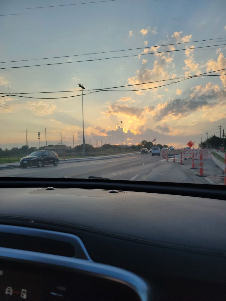 Dramatic sunrise from behind clouds, over a very mundane street with traffic cones. Photo taken from passenger side view inside a car 