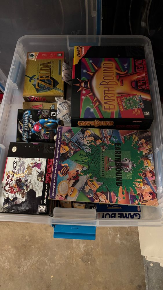 A plastic bin full of video game boxes and items.  Visible are boxes for the SNES games Earthbound and Chrono Trigger, GBA games Metroid Fusion and Minish Cap, N64 game Ocarina of Time (Special Edition), and a well used player’s guide for Earthbound.  Strewn about within are packets of desiccants.