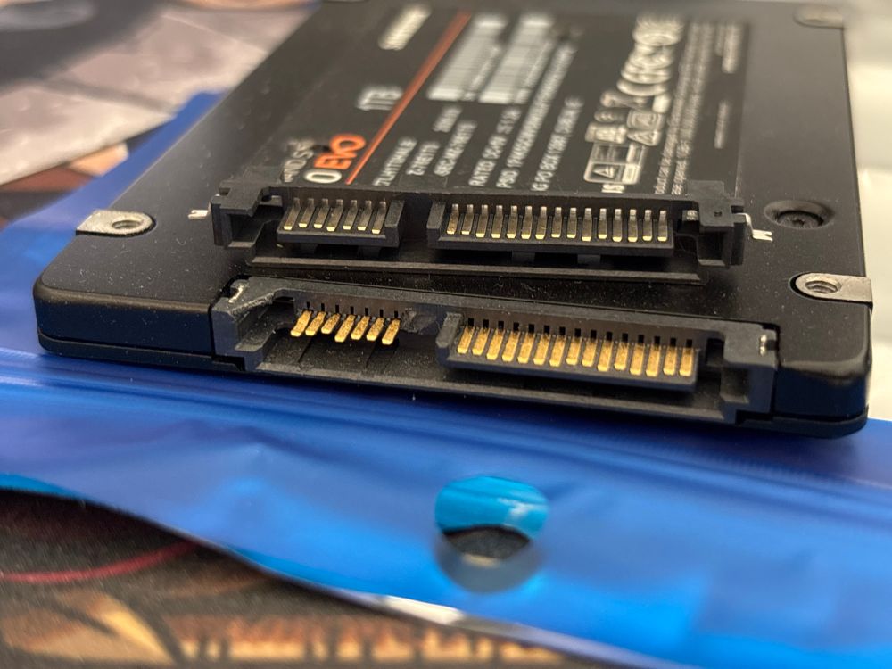 A side view of the SSD with the old broken connector below, and an all new replacement connector with the missing plastic support on top of it.