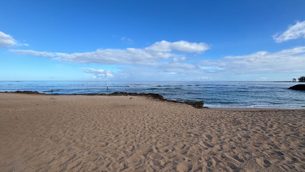 A wide angle picture of a beachfront, showing a partially rocky shoreline.  A few surfers are visible on the water but the waves are mostly calm.