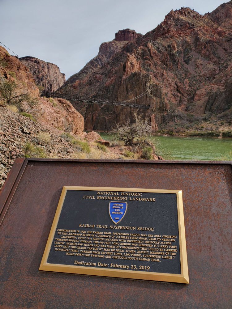 The Black Suspension Bridge, a dark structure that blends into the shadowed red rocks behind it, crosses the green waters of the Colorado River at the end of the South Kaibab Trail into the Grand Canyon.  This National Historic Civil Engineering Landmark, a plaque about which is in the foreground, was completed in 1928.  In the era before helicopters, all of the beams, cables, manpower, and supplies had to be carried down the 7-mile trail.  Much of this back-breaking labor was done by members of the Havasupai tribe.  Today the bridge still serves as the crossing point from the South Kaibab Trail to Bright Angel Campground and Phantom Ranch.