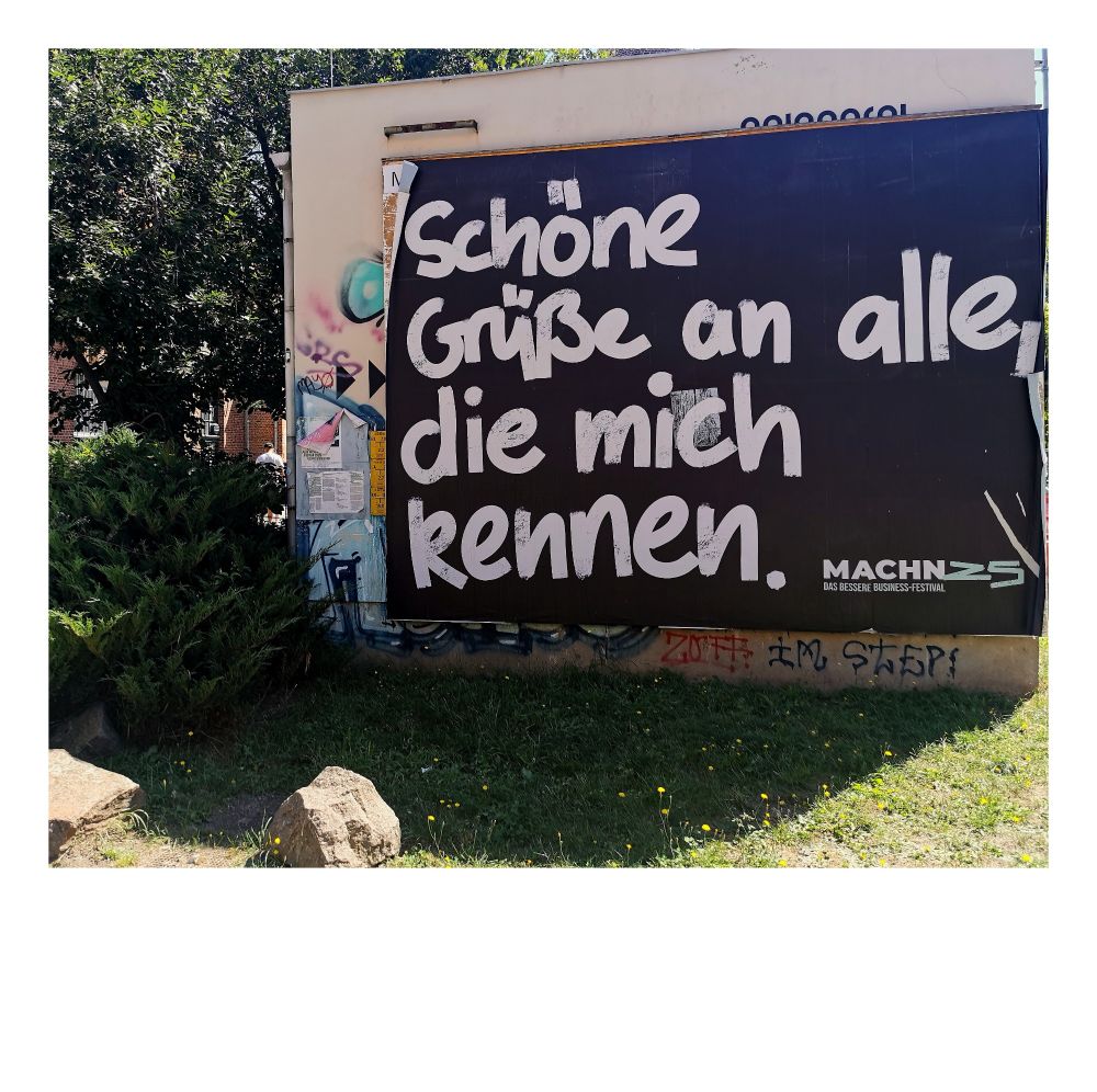 Das Bild zeigt ein großes, braunes Plakat mit der Aufschrift: „Schöne Grüße an alle, die mich kennen.“ Dieses Plakat ist an einer Wand angebracht, umgeben von etwas Grün, möglicherweise in einer städtischen Umgebung. Es vermittelt eine freundliche Botschaft an die Menschen, die die Person kennen, die es angebracht hat.