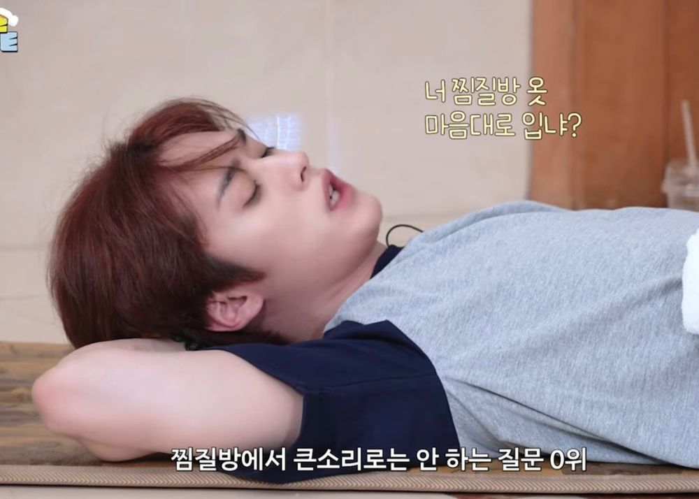 Minho in a grey t-shirt with blue sleeves looking down, laying down, his arm behind his head, lips parted