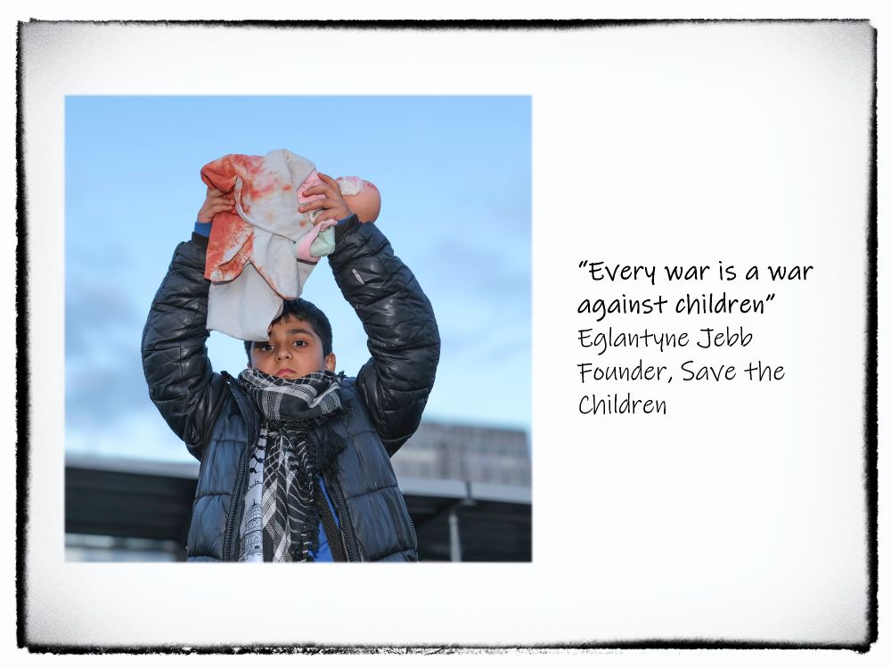 Photograph of a child holding up an effigy of a dead baby, wrapped in cloth and covered in blood with the following words to the right:
"Every war is a war
 against children"
 Eglantyne Jebb
 Founder, Save the
 Children