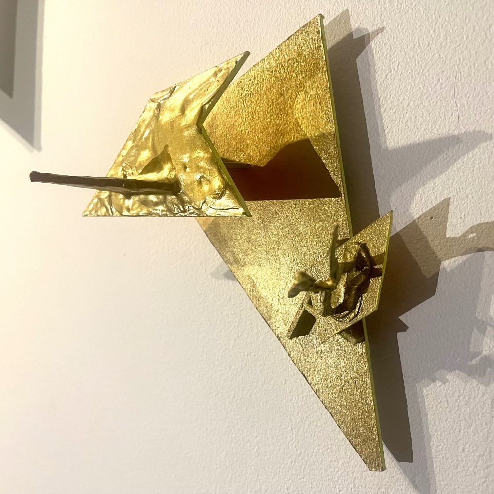 A small wall sculpture made of cardboard, with different levels of different shapes. On one of the parts there is a small toy soldier holding a gun. All is painted gold, and the edges of the shapes are painted reflex yellow. There is an orange reflexion on the wall. The overall size is 17×16 cm.