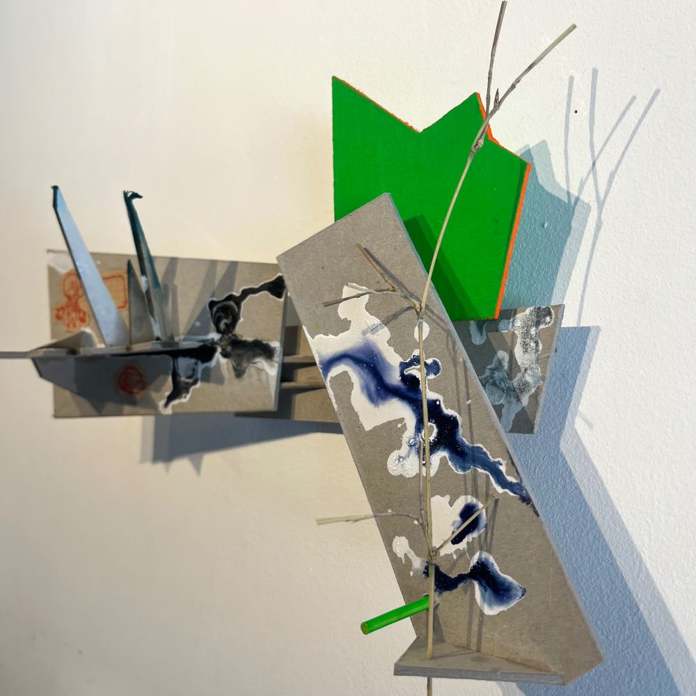 A small wall sculpture made of cardboard, with little  pikes of cardboard, and splashes of dark and white ink. A part of it is painted in reflex green. A tree chunk is planted on the right. Some little drawings are glued : a man with an oversized mask speaking an empty speech bubble, a house with puffs of smoke, a rock.
