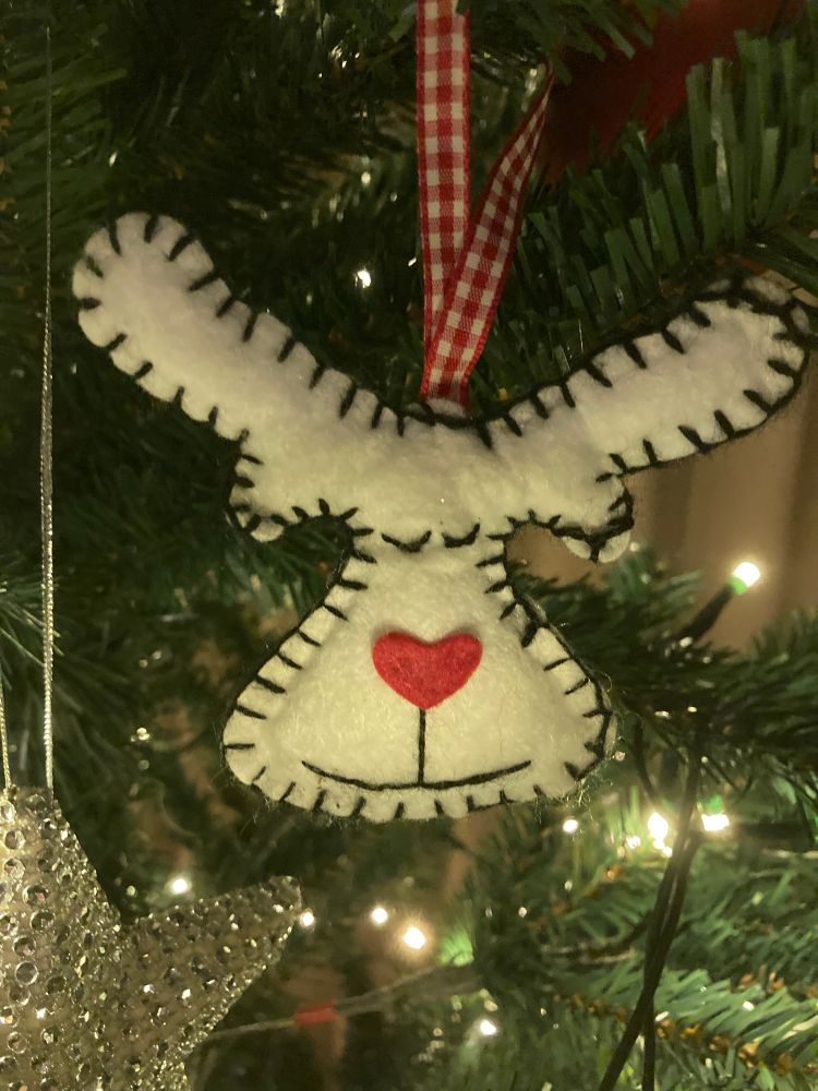 Viltfiguur in de vorm van een rendierhoofd, gemaakt van wit vilt met zwarte stiksels langs de randen. Het figuurtje heeft een rood hartje als neus, twee zwarte lijnen als mond en een rood-wit geblokt lintje waarmee het aan een kerstboom hangt. Op de achtergrond zijn groene dennentakken, kerstverlichting en een zilverkleurig, glinsterend sterornament te zien.