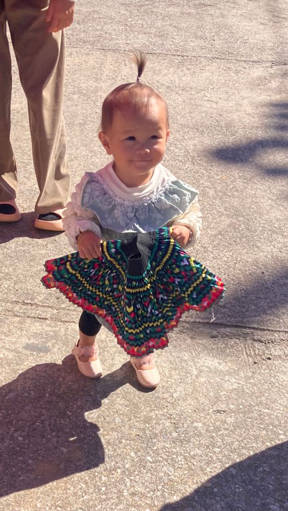 Een klein meisje, staand op een zonnige betonnen ondergrond. Ze heeft een paar lichte schoenen aan en draagt donkere leggings. Haar bovenkleding bestaat uit een witte blouse met kant en een lichtblauw schortje met een patroon. Ze houdt een kleurrijke rok of kleedje vast, met zigzag patronen in groen, geel, rood, blauw en zwart. Ze heeft een klein staartje boven op haar hoofd gebonden. Op de achtergrond zijn de benen te zien van een volwassene in beige broek en zwarte schoenen. De zon werpt schaduwen op de grond.