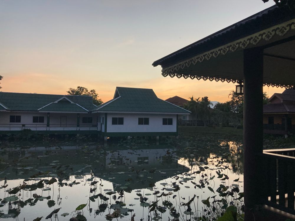 Huisjes met groene daken op palen boven een vijver vol met waterplanten, bij zonsondergang. De lucht is zacht oranje-blauw gekleurd. Rechts is een deel van een overkapping met decoratieve randen zichtbaar. Op de achtergrond staan bomen en andere huizen. Het geheel straalt rust en een tropische sfeer uit.