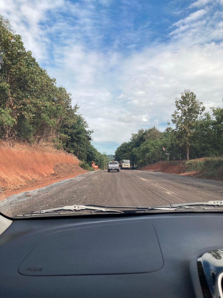 Weg in aanbouw gezien vanuit een auto. Voor het raam van de auto zie je een brede tweebaansweg die nog niet volledig geasfalteerd is, met losse steentjes op het wegdek. Aan beide kanten van de weg staan bomen en struiken, met links een stuk omgewoelde rode aarde. In de verte rijden enkele voertuigen, waaronder een witte auto en een vrachtwagen. De lucht is deels bewolkt maar met blauwe stukken zichtbaar.