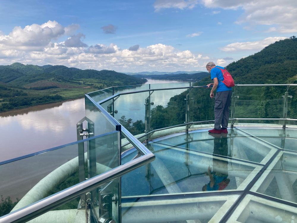 Uitkijkplatform met glazen vloer, gebouwd boven een rivier en uitkijkend op een weids, groen berglandschap. Je ziet een oudere man met een rode rugzak naar beneden kijken terwijl hij op het glazen platform staat. De rivier stroomt rustig en slingert door het landschap. De lucht is lichtblauw met veel witte wolken en het uitzicht is panoramisch: heuvels en bossen zover je kunt kijken. Het lijkt een populaire plek voor toeristen om van het uitzicht te genieten.