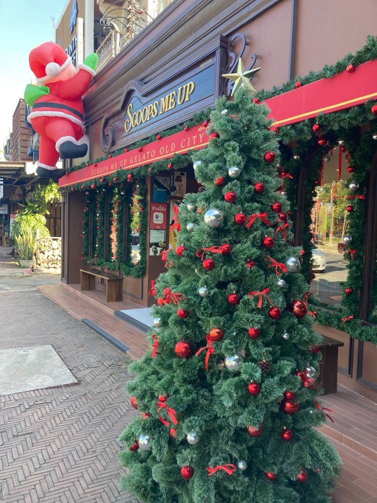 Kerstboom versierd met rode en zilveren ballen, rode strikken en een ster bovenop, die buiten voor een ijssalon staat met de naam "Scoops Me Up". Aan het gebouw hangt een grote opblaasbare kerstman die lijkt te klimmen. De gevel is verder versierd met groene dennentakken en rode linten, wat een feestelijke kerstsfeer creëert.