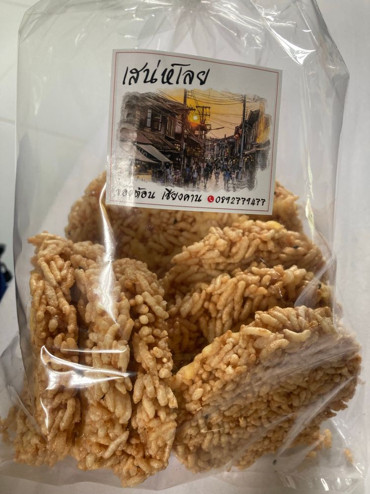 Zak met Thaise rijstsnack. In een doorzichtige plastic zak zitten ronde, krokante rijstwafels die aan elkaar geplakt lijken door een zoete siroop. Op het etiket staat tekst in het Thais, met daaronder een tekening van een straatbeeld met mensen en traditionele gebouwen. De snack ziet eruit als een typische Aziatische zoete rijstkoek.