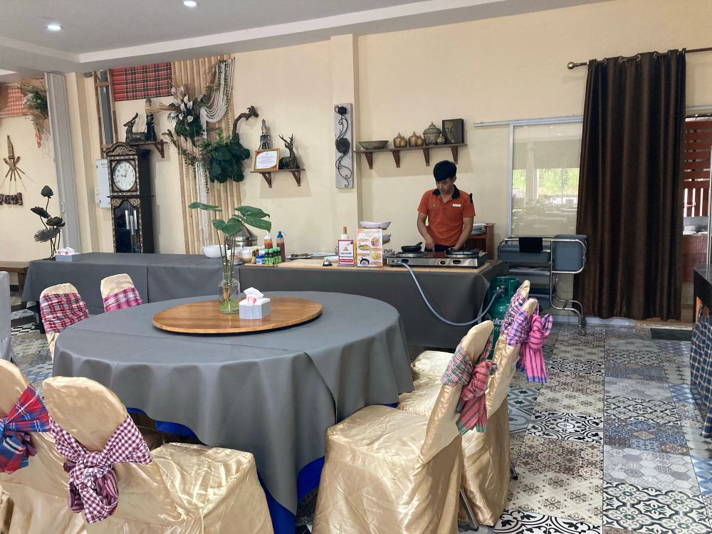 Omelet of spiegel ei vers gemaakt.

Restaurantinterieur met ronde tafel in het midden, bedekt met een grijs tafelkleed en een draaiplateau erop. De stoelen zijn bekleed met goudkleurige stoelhoezen en versierd met geruite linten in verschillende kleuren. Op de achtergrond staan decoratieve voorwerpen, een grote staande klok, en planten. Aan de rechterkant staat een man in een oranje shirt achter een buffet of kookstation, waar flessen saus en ingrediënten te zien zijn. De vloer is bedekt met sierlijke, kleurrijke tegels in diverse patronen. Aan de muur hangen schalen, kunstvoorwerpen en een gordijn. De sfeer is huiselijk en feestelijk.