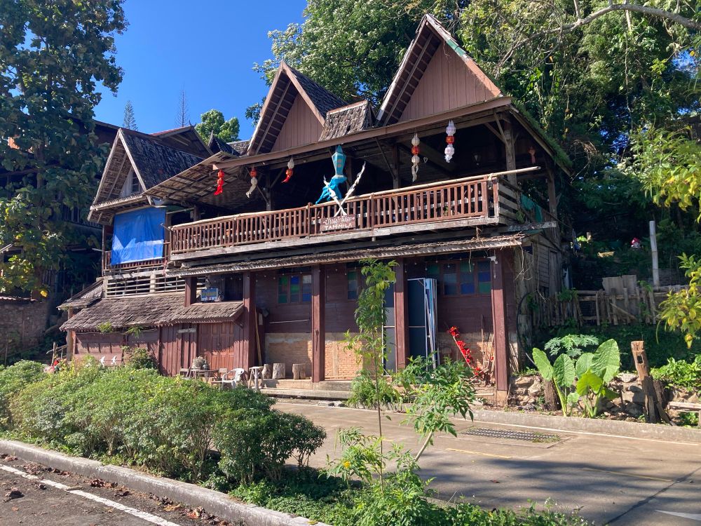 Houten huis in traditionele stijl. Dit huis heeft een hoge, schuine dakconstructie met meerdere punten, en een ruime veranda op de bovenverdieping met een houten balustrade. Op de veranda hangen gekleurde decoraties en lampionnen. De gevel bestaat uit houten planken en ramen met gekleurd glas. Voor het huis staan enkele witte stoelen aan de linkerkant. Rond het huis groeit veel groen, met struiken en grote bladeren, en daarachter staan hoge bomen. De lucht is helderblauw en het geheel heeft een tropische, landelijke uitstraling.