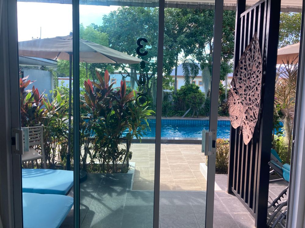 Zicht door een glazen schuifdeur van onze kamer op een zonnige tuin met zwembad. Aan de linkerkant staan twee blauwe ligstoelen naast elkaar, en er is een grote parasol zichtbaar boven wat kleurrijke planten. Rechts hangt een decoratief, uitgesneden kunstwerk in de vorm van een vis aan een zwart hekwerk. Buiten zijn bomen, struiken en planten te zien rondom het blauwe zwembad. Het geheel ademt een ontspannen en tropische sfeer uit.