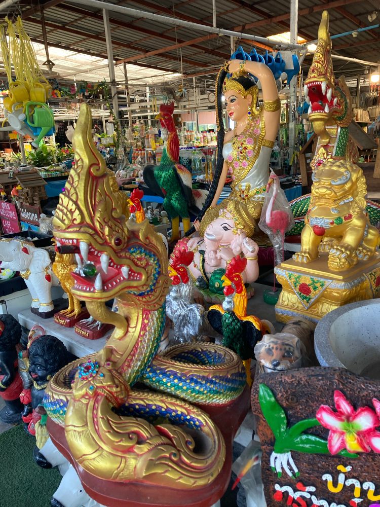 Beeldjes van mythologische wezens en goden. Op de voorgrond staat een groot, felgekleurd standbeeld van een draak of slang, rijkelijk versierd in goud, rood, blauw en groen. Ernaast staan nog meer beelden, waaronder een vrouwelijk figuur in traditionele kledij, een mythische leeuw en een paar hanen. Je ziet ook andere beelden in verschillende kleuren en stijlen op de achtergrond. Het geheel lijkt te staan in een overdekte markt of winkel waar decoratieve beelden en tuinornamenten worden verkocht. Overal in het beeld zie je veel kleur en details.