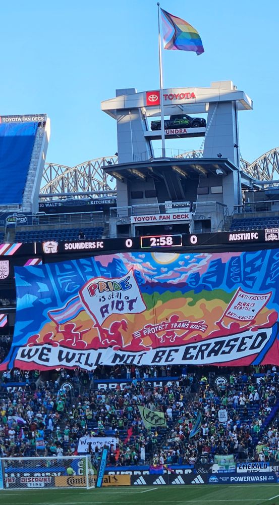 Pride Night at the Seattle Sounders game. The supporters section raises a huge banner saying "PRIDE is still a RIOT. Anti-fascist, anti-racist, always Seattle. Protect Trans Kids. WE WILL NOT BE ERASED". The trans rainbow flag waves from the stadium flagpole.