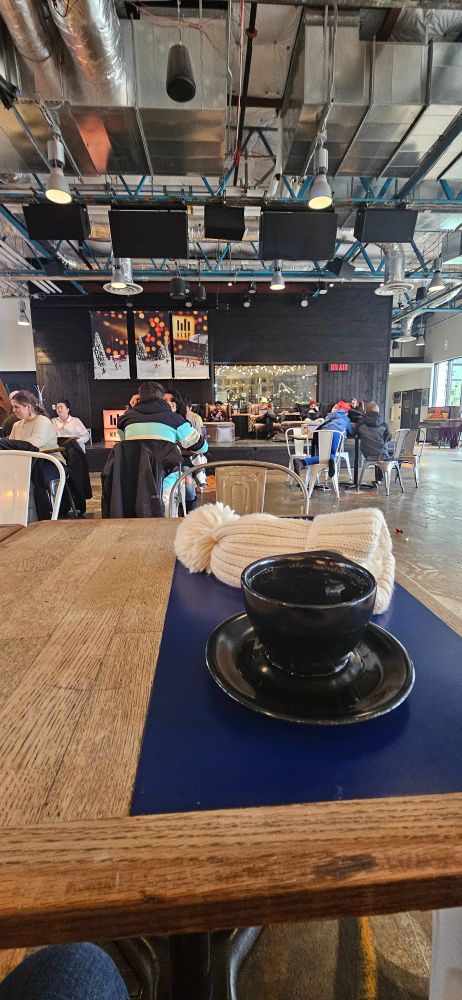 Sitting in the KEXP Gathering Lounge with other people of all ages chat and relax. A steaming cup of black coffee is in the foreground with a white winter hat, recently removed, behind