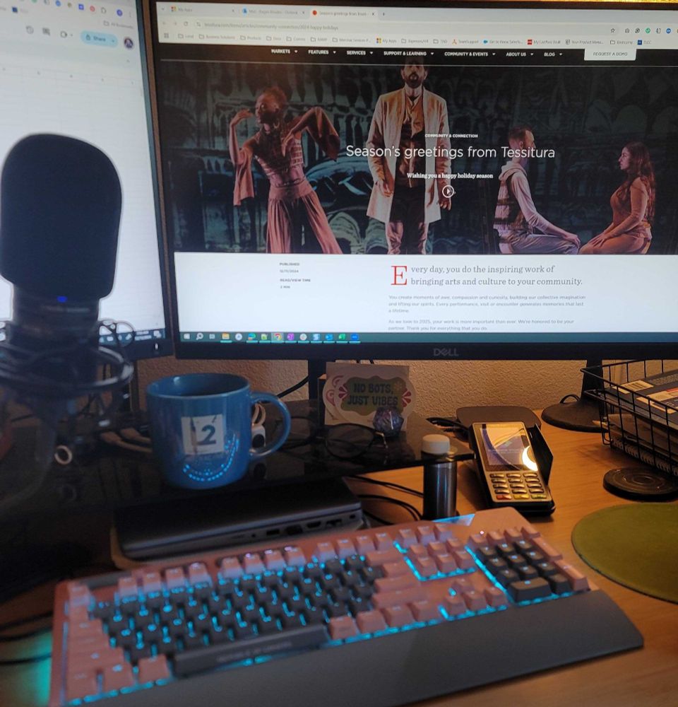 A desk setup showing a computer monitor displaying a website for Tessitura with a holiday greeting message and theatrical performance images. The desk has a backlit pink mechanical keyboard with blue lighting, a blue mug with the company logo L2 printed on it, a microphone on a stand, and various desk accessories including glasses and a small sign that reads 'NO BOTS JUST VIBES'. 