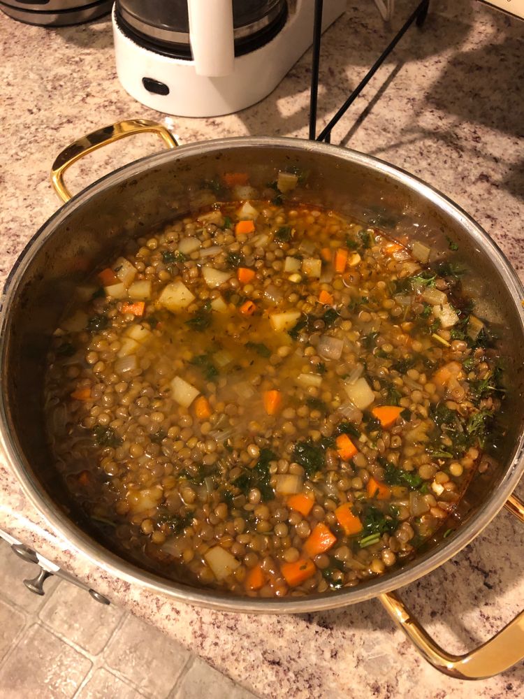 A pot of lentil soup. Brown lentils, carrots, parsley, and potatoes in a steel pot with gold colored handles on a faux granite counter. White coffee maker in the top left corner. 