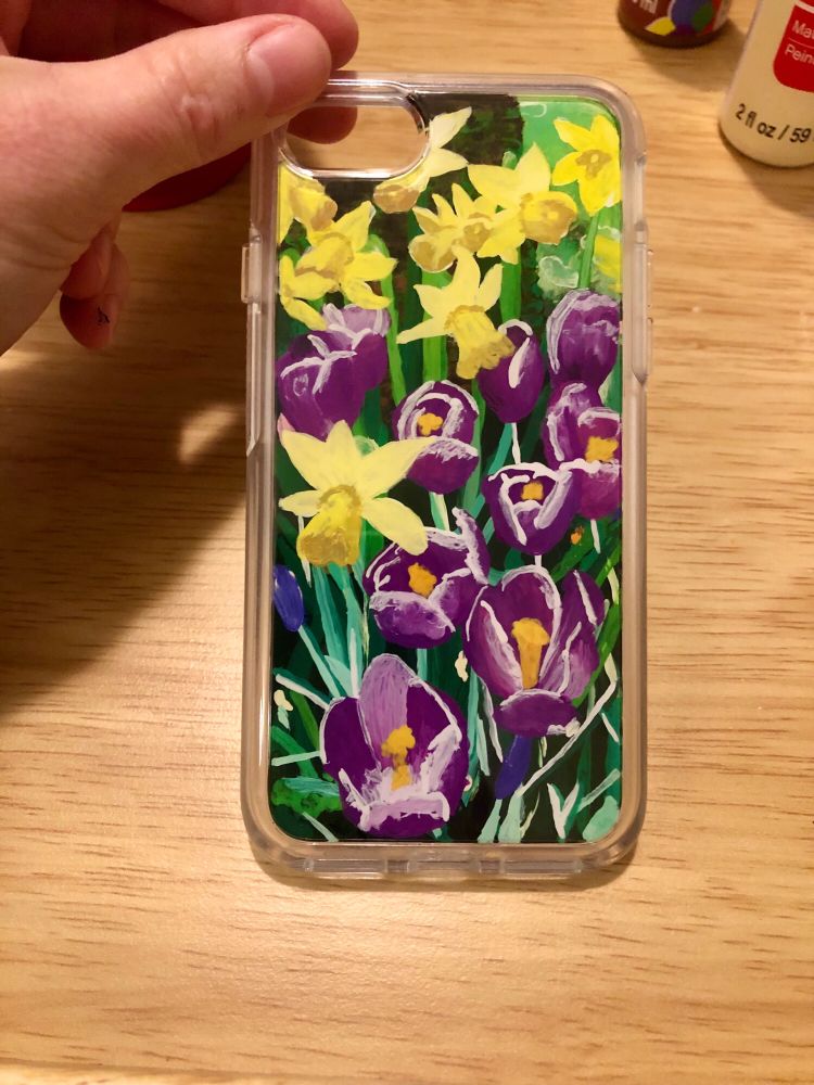 A phone case is held at an angle by a hand on a wooden surface. The top right corner shows two bottles of paint, white and purple, that sit on the surface. The entire back panel of the case is painted. The painting depicts purple crocuses in the bottom, right, and center, and yellow daffodils at the top, left and center. Blades of grass surround the flowers, and a tree trunk, almost completely hidden by the flowers, stands in the top left corner.