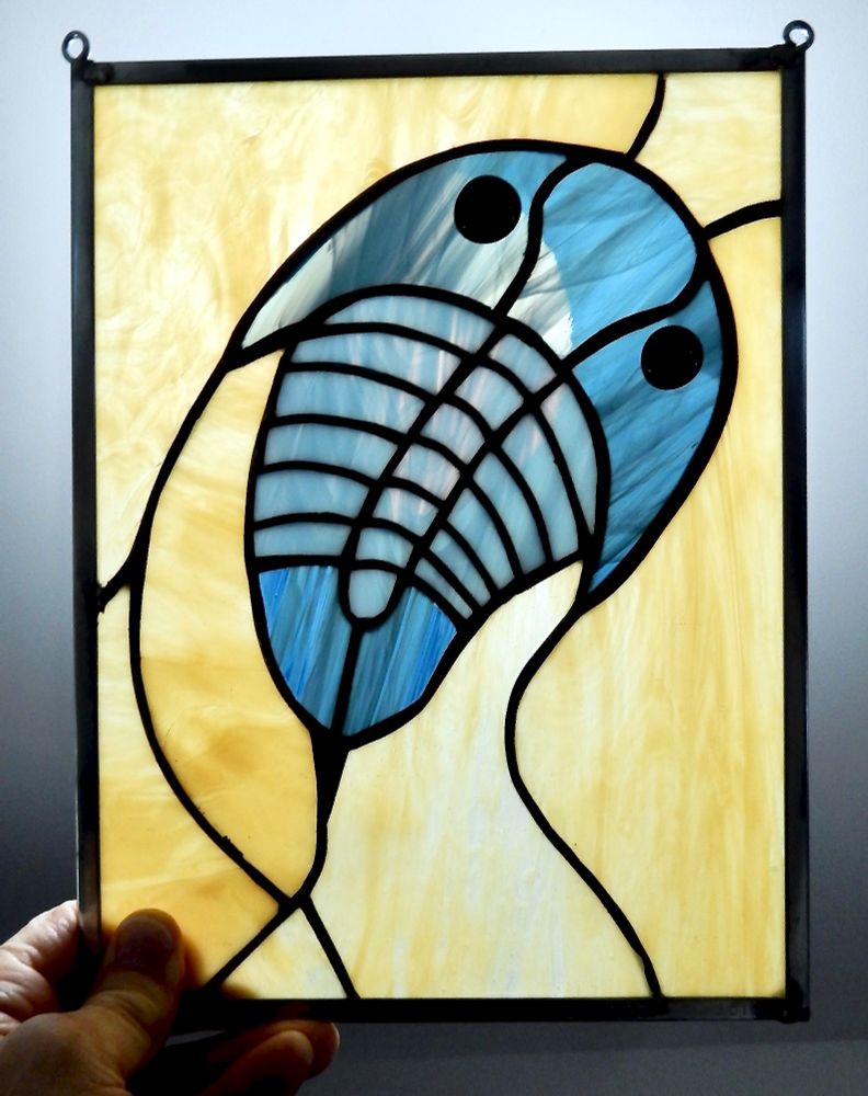 A human hand holds up a small, back-lit, stained glass panel depicting a graceful swimming trilobite in pale blue tones. It is surrounded by honey-coloured glass in a narrow metal frame. 
