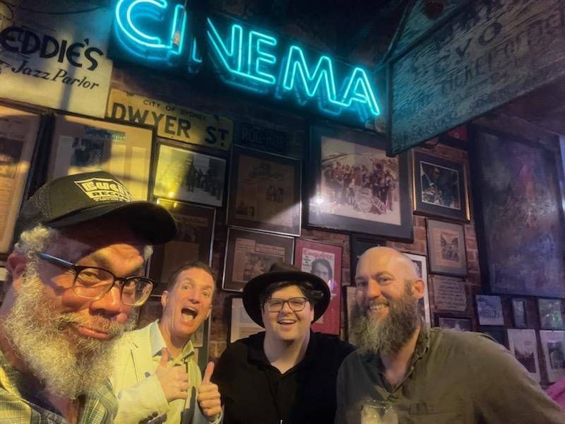Four men smiling at the camera, in front of a wall of photos.  A blueish neon sign at the top says CINEMA.