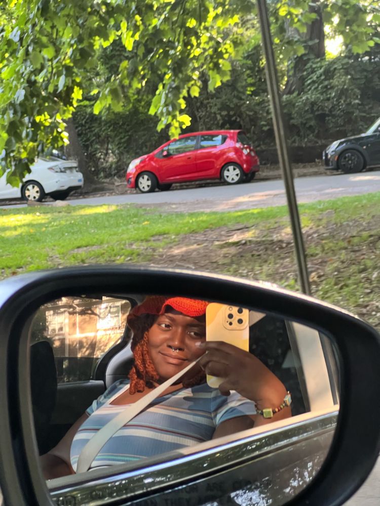 Me (Niya) wearing an orange hat with golden brown locs. I’m holding my phone in a yellow phone case, and taking the picture through the car window. My seatbelt is on, I’m wearing a blue shirt with white, red, green, and yellow stripes. A smile flashes across my face.