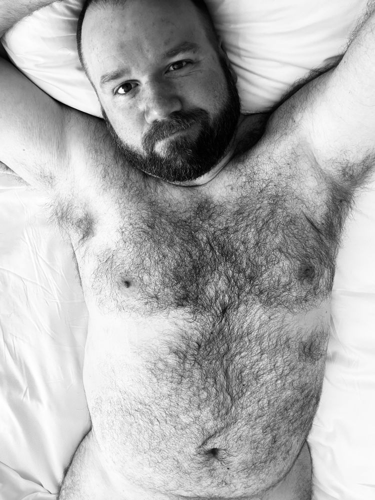 A hair person laying naked on a bed looking up at the camera. Visible is their torso and head.