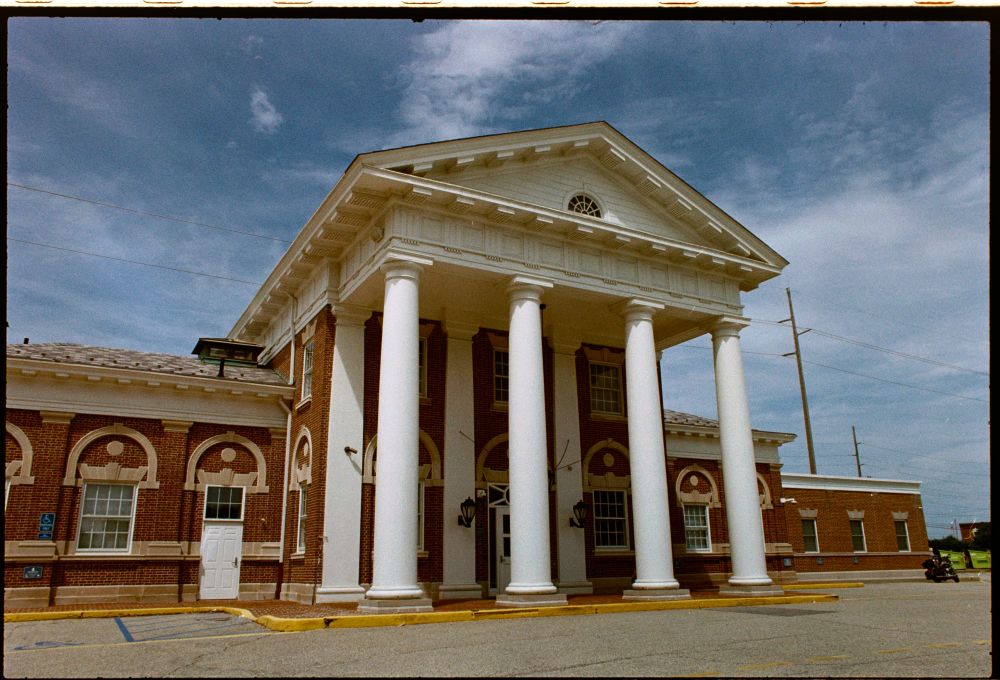 A color film photograph of the former Pennsylvania Station in Dover, DE. SAH Archipedia sez: "Railroad stations once served as triumphal portals to American towns and cities, as this temple-form building recalls. Its Doric portico of wood is heroically massive, as are its wide eaves with mutules. It is reminiscent of Thomas Jefferson's Pavilions at the University of Virginia. The brick building's facade is composed of a series of pilasters and relieving arches under a hipped roof. Massey, a lawyer for the Pennsylvania Railroad, convinced the company to replace the original Italianate station built by the Delaware Railroad (1853–1860), the coming of which had invigorated the southern part of the state."