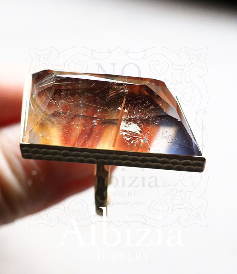 ピクチャーメノウとルチルクォーツのダブレットストーンのリング。変形の長い菱形、ランダムカット、覆輪留め。地金はK18YG。リングを手に持って上向にしやや上から撮影した写真です。金色の地金に青とオレンジと透明の石です。