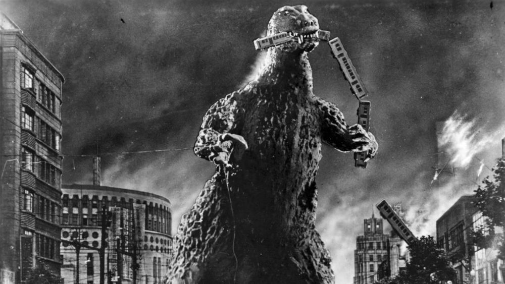 Godzilla monster towering over a burning city while holding and eating some train cars.