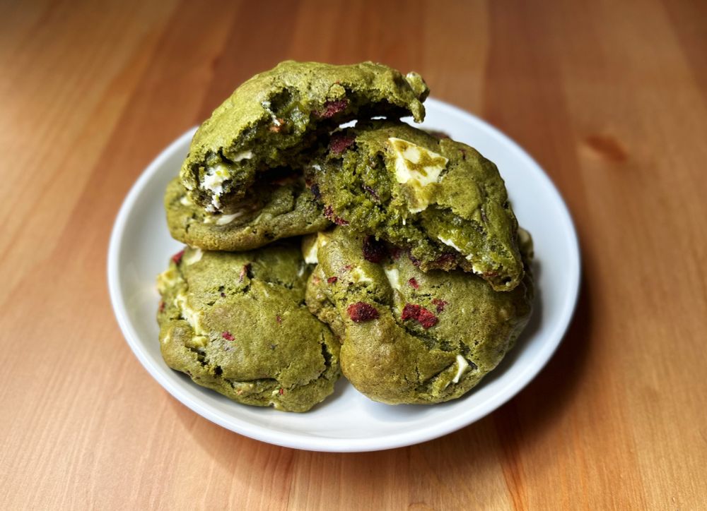 Four matcha cookies, baked with freezer dried strawberries and white chocolate chunks, are stacked on a white plate. The top cookie has been broken in half to let the viewer see the inside. Yes, the freezer dried strawberries did burn a bit. But they still taste great!