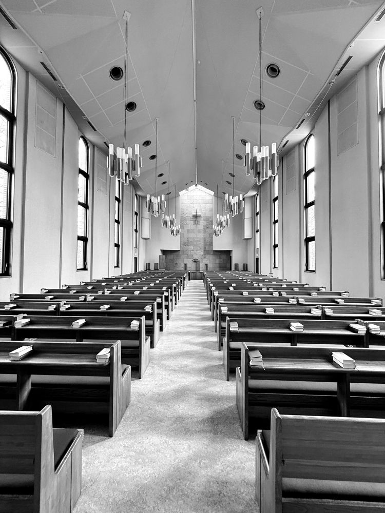 A spacious, modern interior of a chapel featuring wooden pews lined up in rows, with a minimalist altar at the far end. The ceiling is high with angular designs, and there are large windows letting in natural light. Several contemporary light fixtures hang from the ceiling.