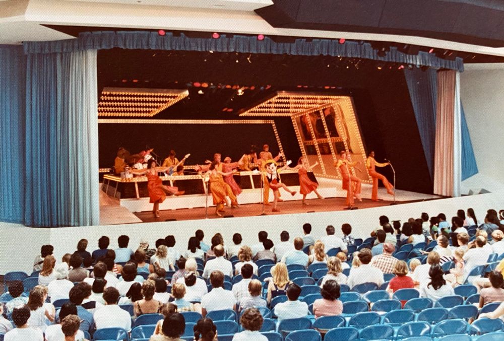 A colorful performance on stage featuring dancers in orange costumes, accompanied by a live band. The audience, seated in blue chairs, watches the show in what appears to be a theater setting.
