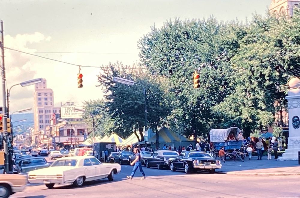 A busy urban intersection from the past, featuring classic cars and a pedestrian crossing the street. A large building is visible in the background with a sign, and tents are set up nearby, indicating a possible fair or market. Lush trees line the street.