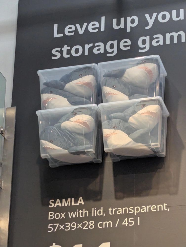 a display, presumably at ikea, reads "level up you[r] storage gam[e]". attached to the wall are four clear plastic storage tubs each containing two stuffed sharks. below the produced is described as a samla, box with lid, transparent