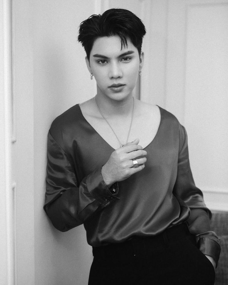 divesth @ instagram | Black and white photo of Fort in a room with white paneled walls, an armchair just visible behind him. He's leaning one against the wall with the one hand in his pocket, the other casually resting over his chest, and a neutral expression. He's wearing black dress pants, a satiny top with long sleeves and a wide, low and rounded neckline, and a necklace with a pendant

#fortfts