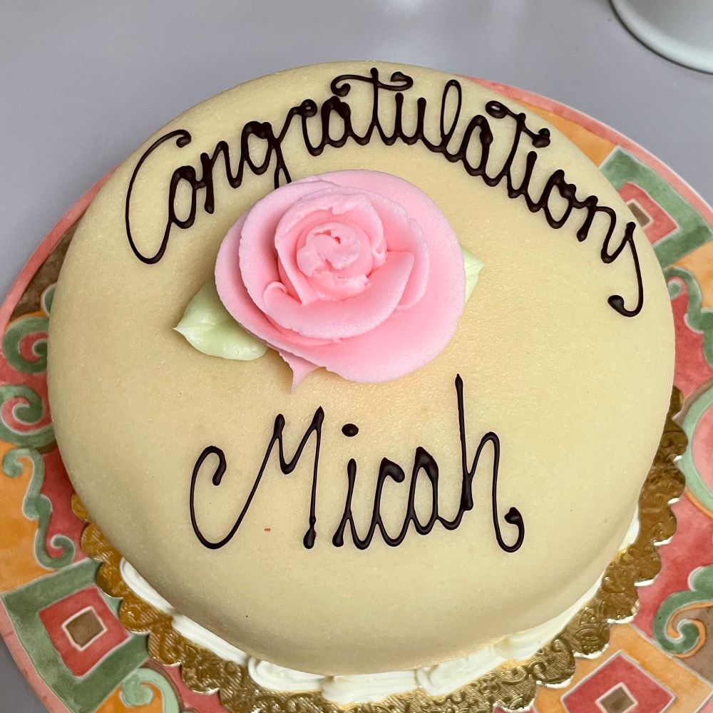 A cream colored princess cake with a pink buttercream rose on top and chocolate lettering reading "congratulations Micah."