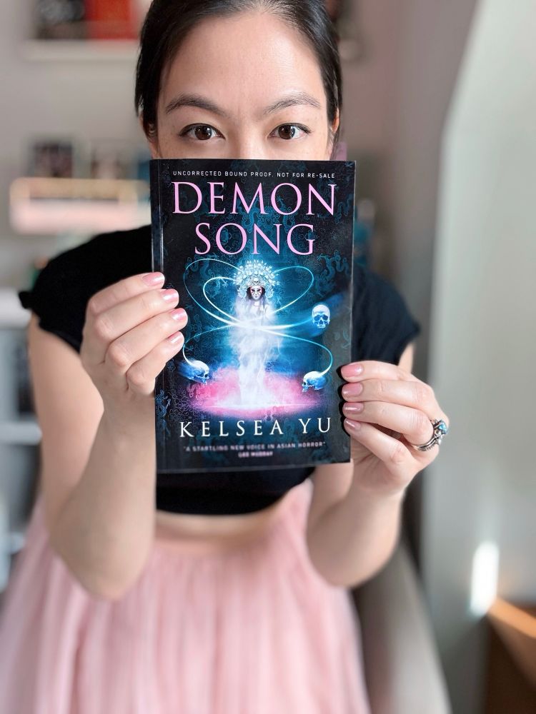 Photo of a Chinese American woman holding a copy of Demon Song by Kelsea Yu in front of her face, with her eyes peeking out from above. She wears a black shirt and pink tulle skirt, and there are books and bookshelves in the background.