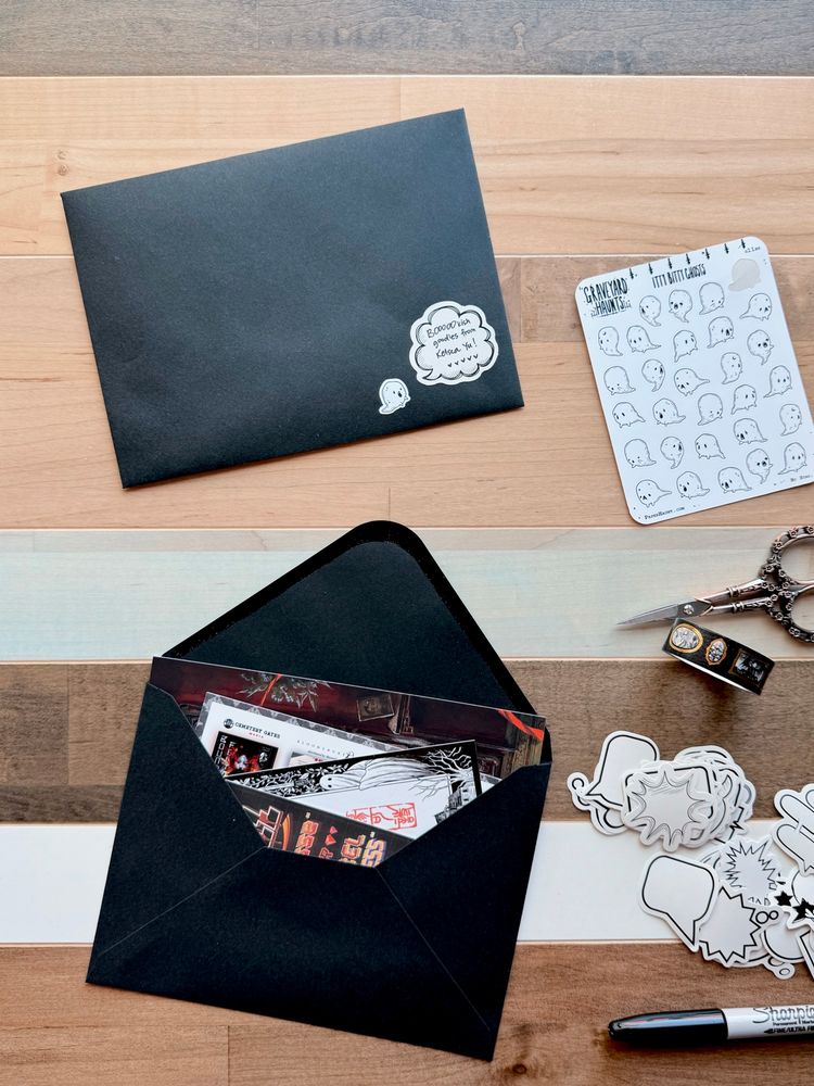 Open envelope showing a sneak peek of several items: art print, postcard, bookplate, bookmark, on a table surrounded by art supplies