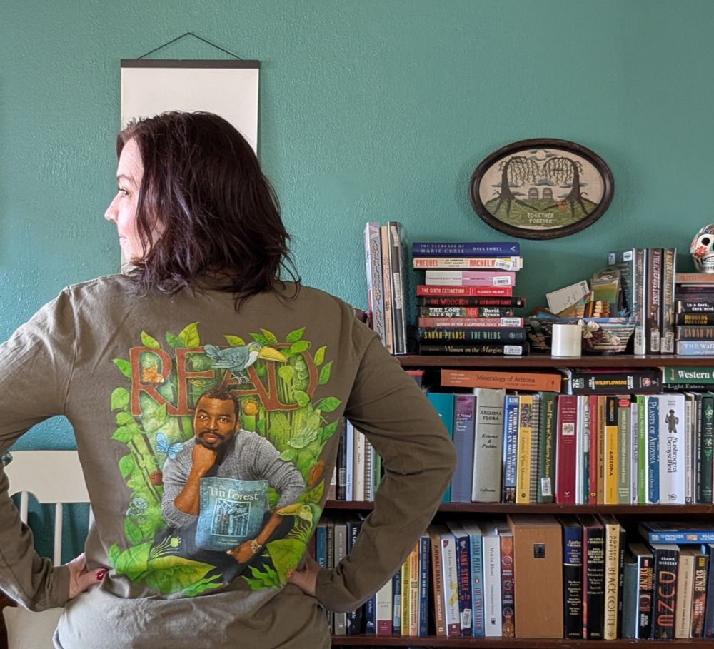A woman stands before a bookshelf with her back turned and hands on hips. She is wearing a t-shirt with an image of LeVar Burton holding a book, with the wording "READ."