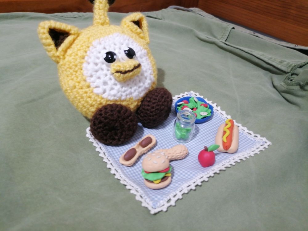 Photograph of a crochet Ouioui doll sitting on a small picnic blanket with plasticine shaped like several food items, such as peanuts, a hotdog, a hamburger, salad, an apple and a jar of pickles. 