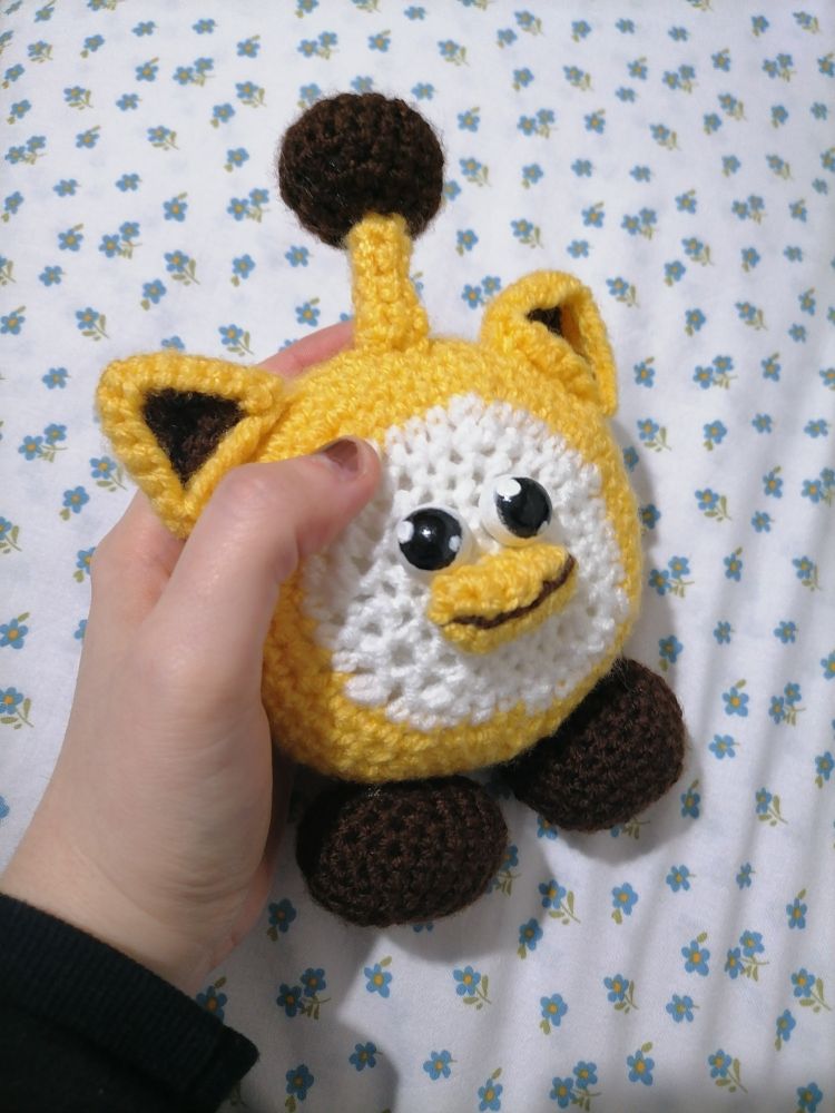 Photograph of a left hand holding a crocheted doll of a Ouioui, a fictional character from the company Itemlabel, that resembles a ball with a beak, cat ears, an antenna and two little feet. 
